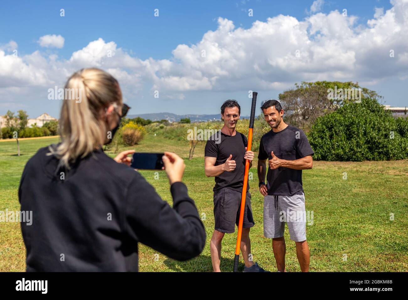 Fitness trainer e un cliente posa per una donna reporter, sparando su uno smartphone. Felice e soddisfatto. Con bastone per ginnastica. Estate in giardino. Foto Stock