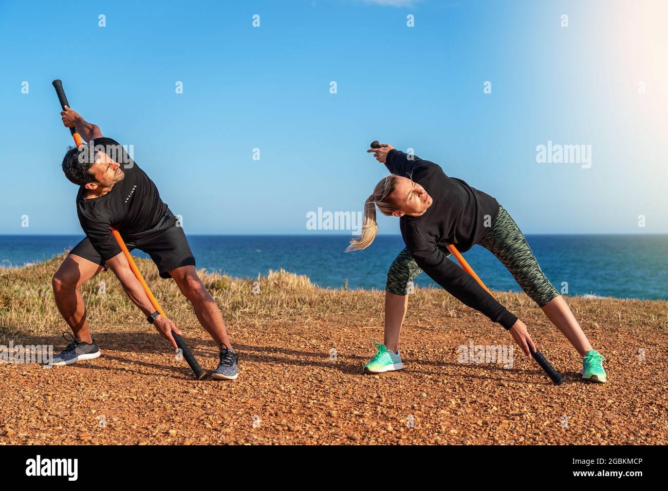 Un uomo e una donna di mezza età sull'oceano stanno facendo un riscaldamento. Lezioni di fitness. Utilizzo di bastoncini da ginnastica per l'allungamento. Giorno di sole. Foto Stock