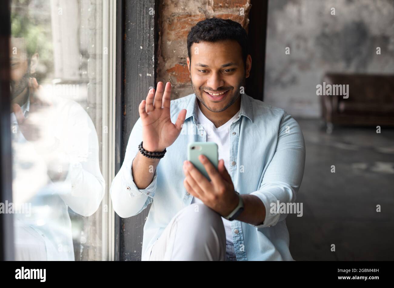 Gioioso uomo indiano che parla con videochiamata sullo smartphone, seduto sul davanzale, ciao in webcam, felice di incontrare amici o familiari a distanza Foto Stock