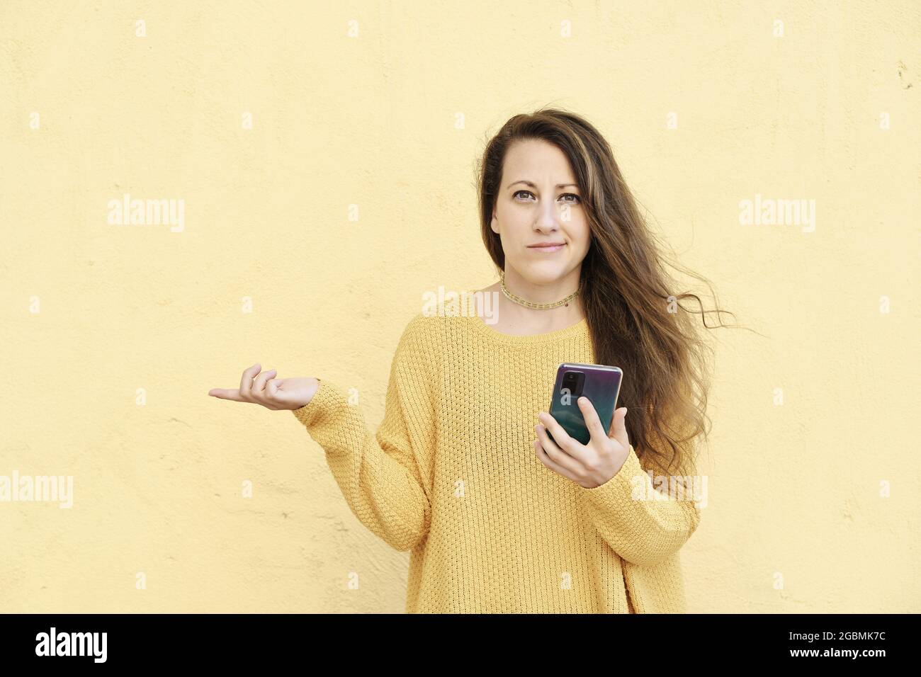 giovane bionda donna perplessa ed esitante con un'espressione divertita usando uno smartphone Foto Stock