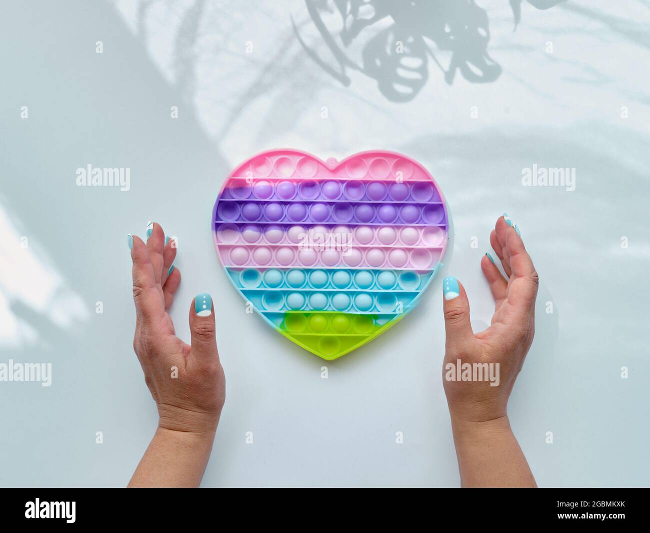 Giocattolo antistress a forma di cuore del papa in mani femminili. Fidget a concavità sensoriale semplice nei colori di LGBT. Sfondo bianco con ombre floreali alla moda Foto Stock