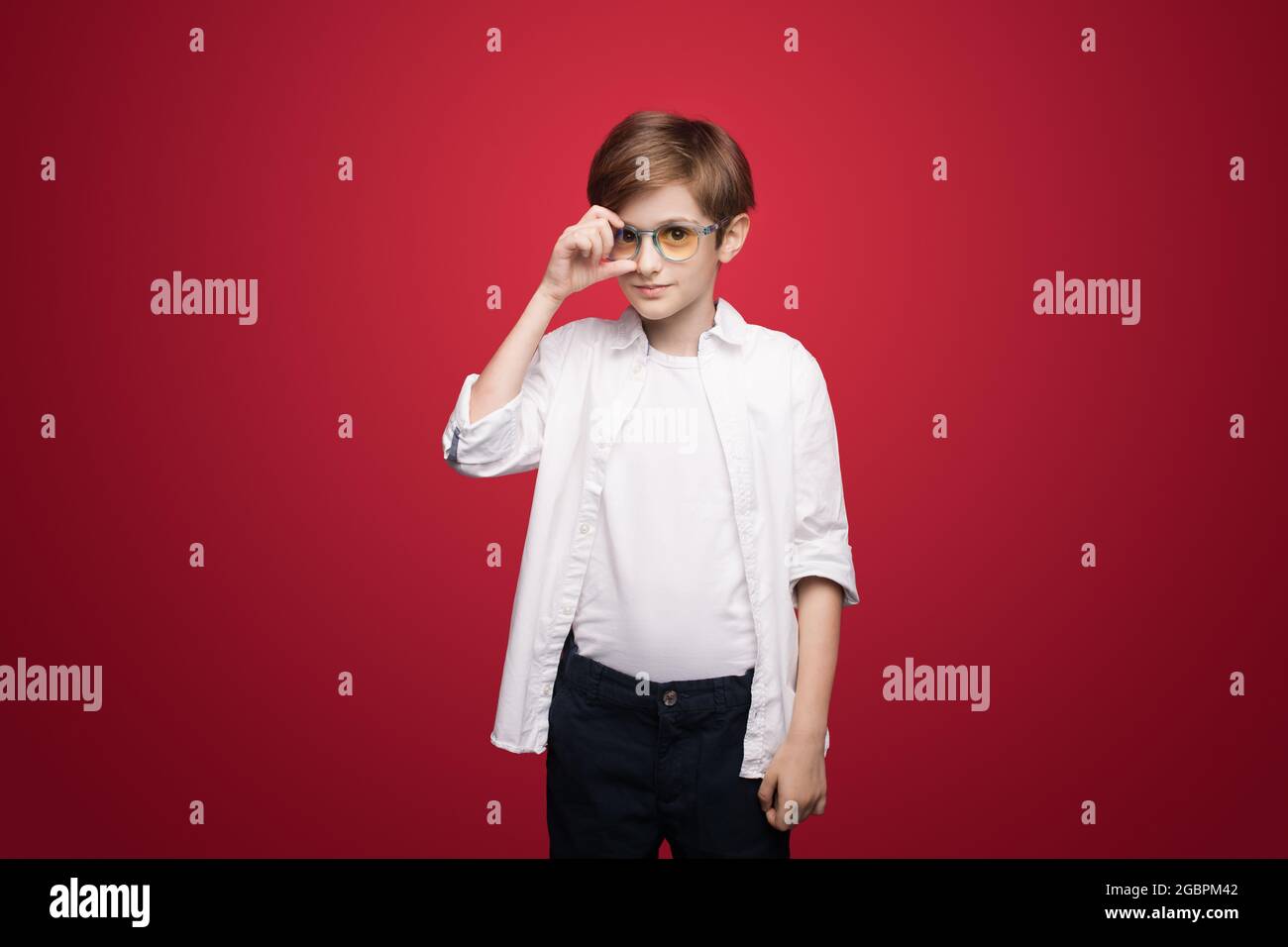 Adorabile ragazzo tocca i suoi occhiali che si posano su una parete rossa dello studio in camicia bianca guardando la macchina fotografica Foto Stock