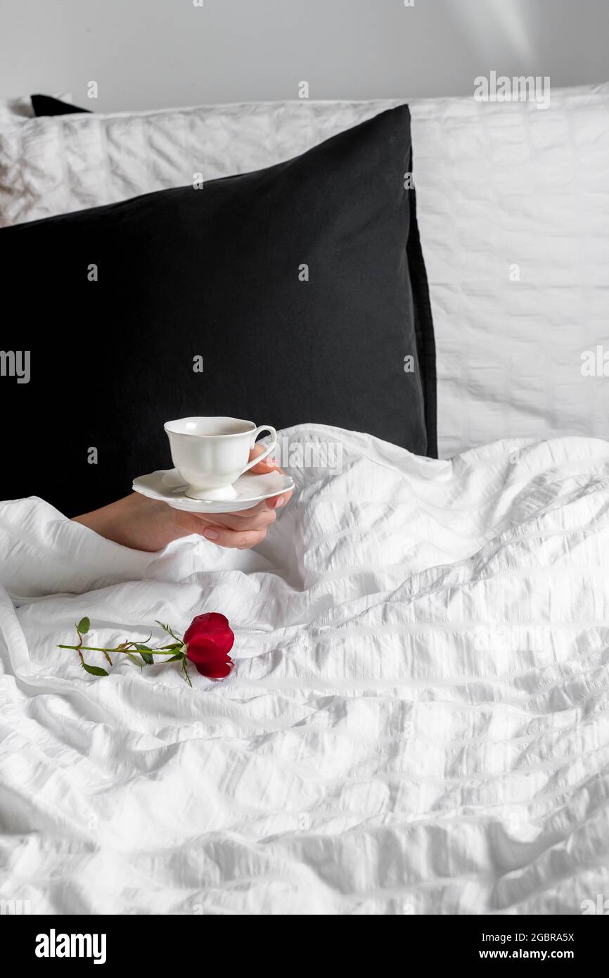 Mano femminile con tazza di caffè e rosa rossa in biancheria da letto bianca Foto Stock