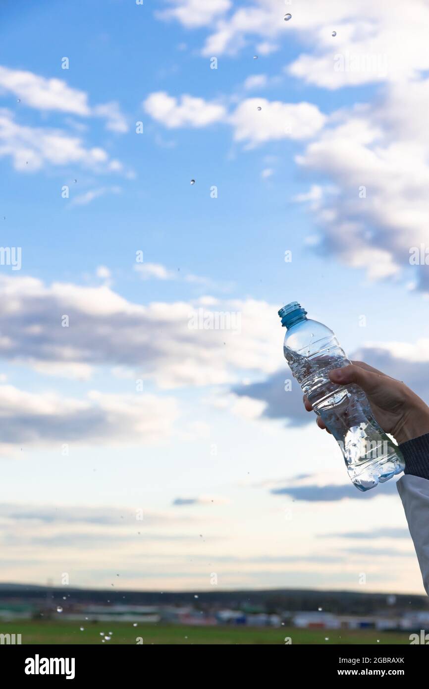 La mano di una donna solleva una bottiglia di acqua minerale pura altamente trasparente. Gli schizzi volano sullo sfondo di un cielo blu con nuvole bianche. S Foto Stock