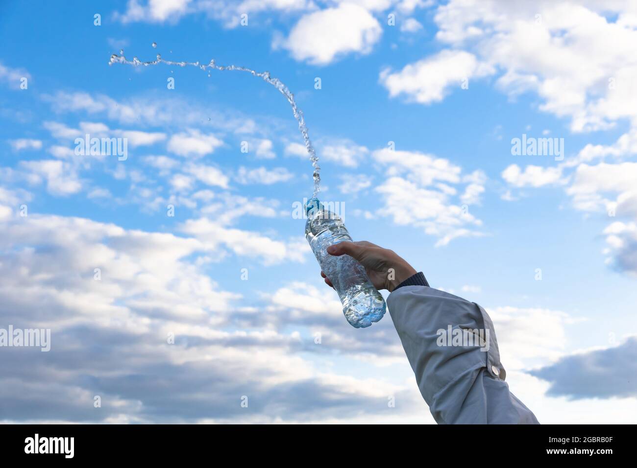 La mano di una donna solleva una bottiglia di acqua minerale pura altamente trasparente. Gli schizzi volano sullo sfondo di un cielo blu con nuvole bianche. S Foto Stock