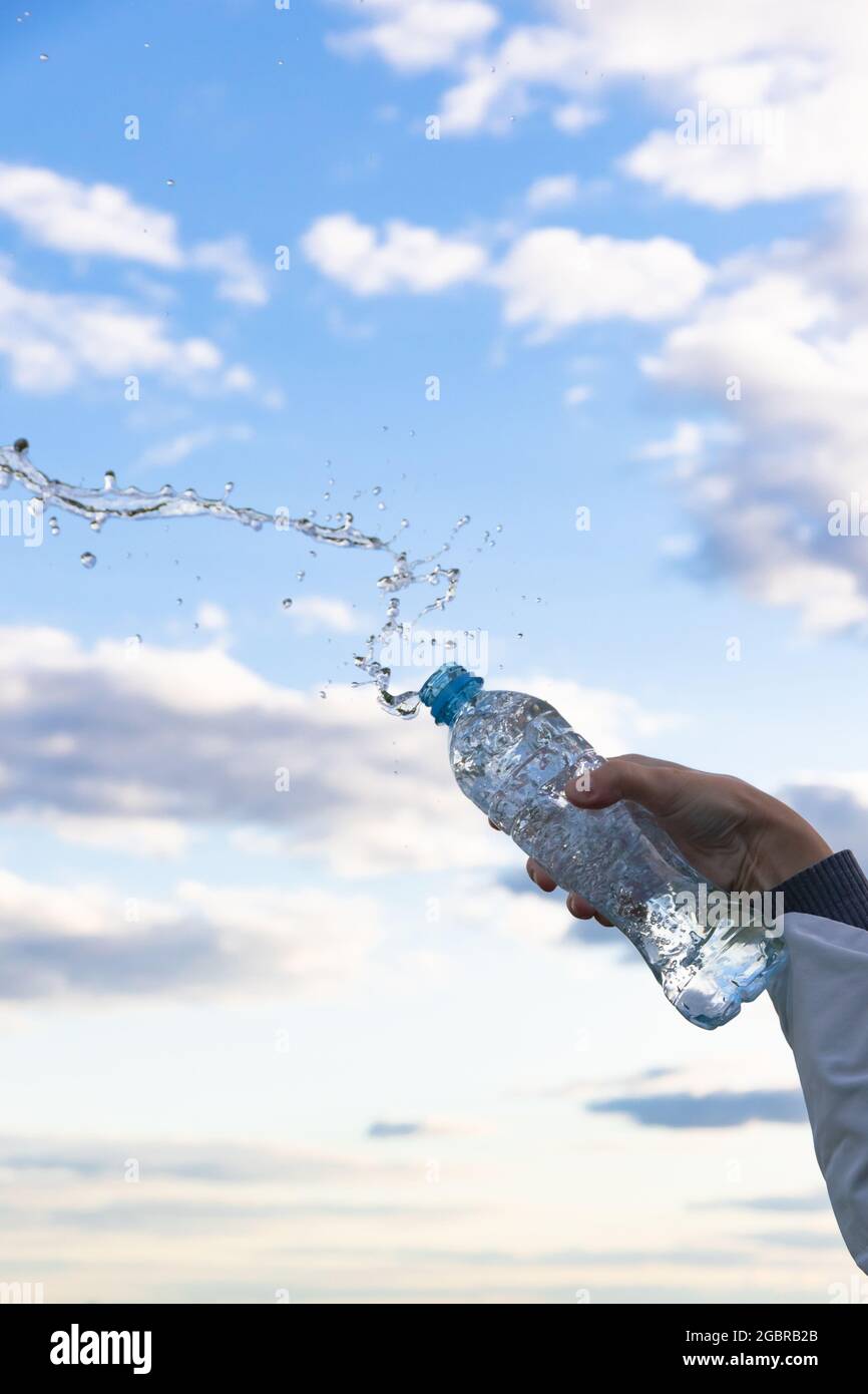 La mano di una donna solleva una bottiglia di acqua minerale pura altamente trasparente. Gli schizzi volano sullo sfondo di un cielo blu con nuvole bianche. S Foto Stock
