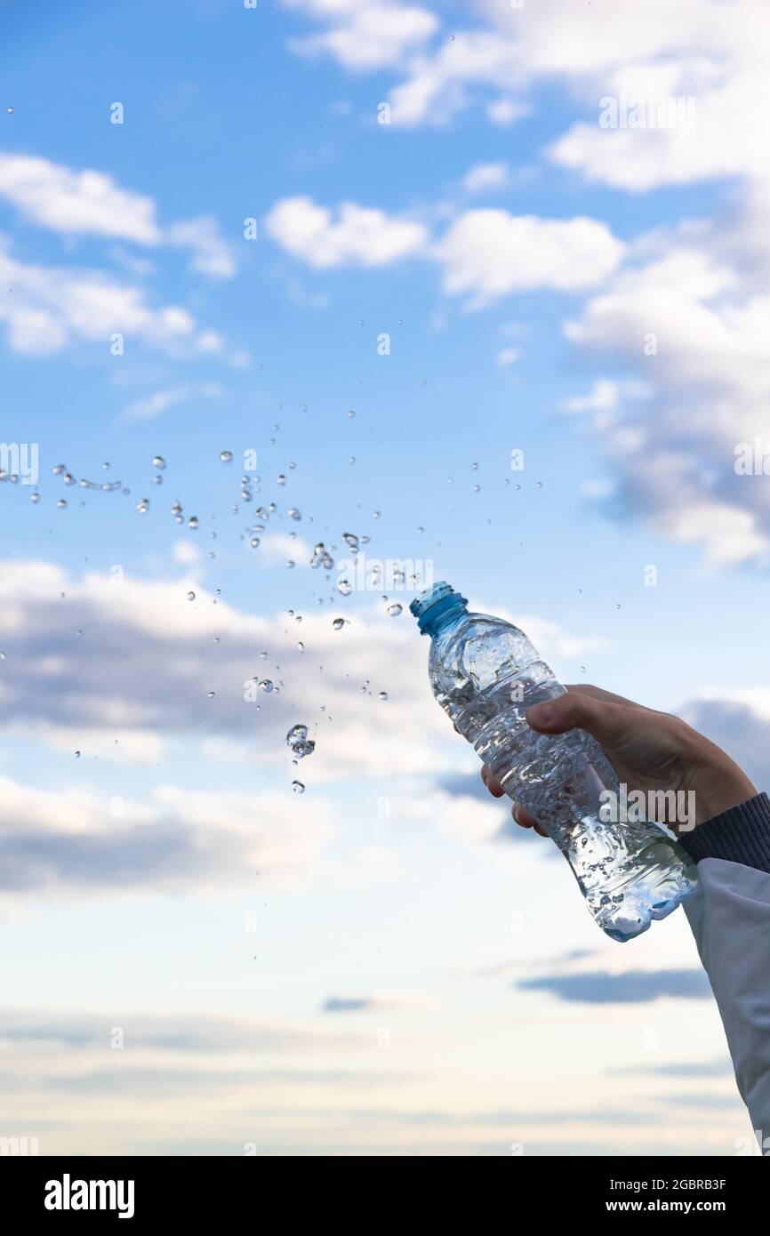 La mano di una donna solleva una bottiglia di acqua minerale pura altamente trasparente. Gli schizzi volano sullo sfondo di un cielo blu con nuvole bianche. S Foto Stock