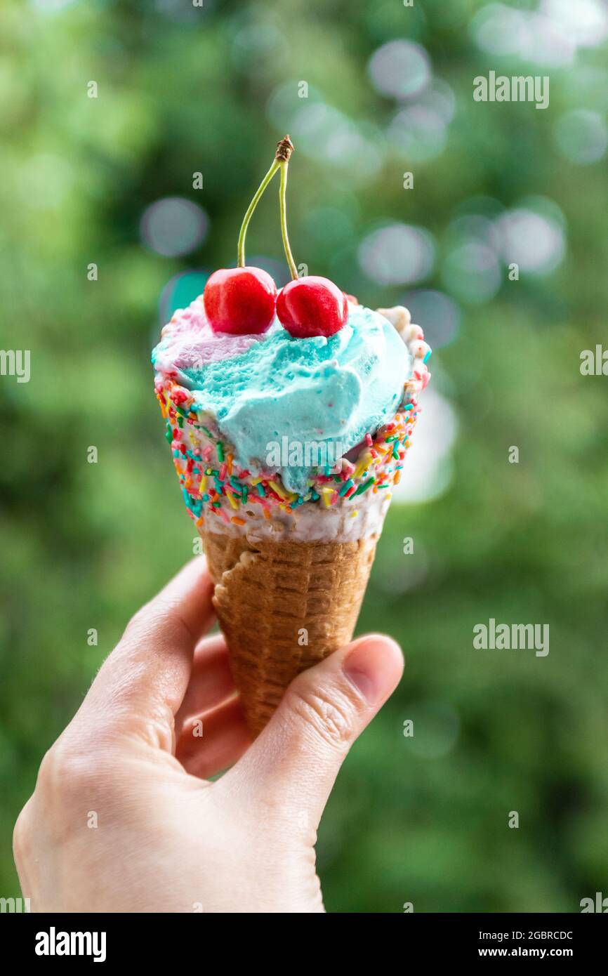La mano femminile tiene il gelato multicolore con le ciliegie in un cono di cialda su sfondo verde Foto Stock