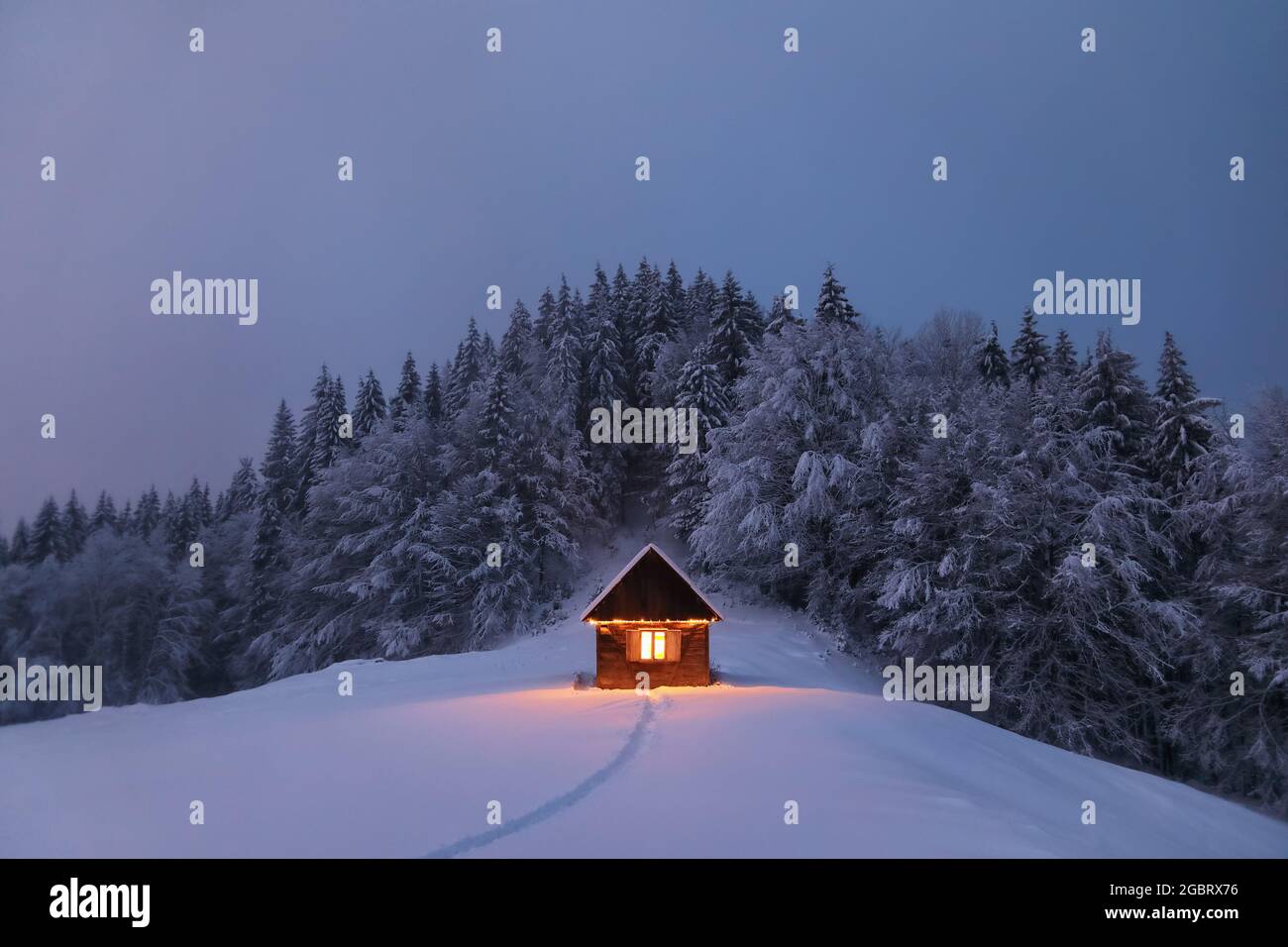 Paesaggio invernale. Notte mistica. Vecchia capanna di legno sul prato coperto di neve. Le lampade illuminano la casa alla sera. Montagne e foreste Foto Stock