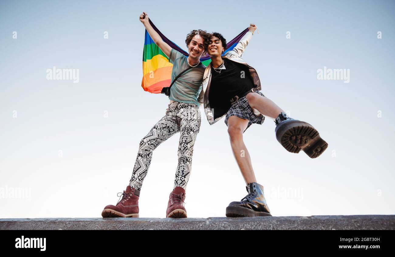 Due giovani uomini che celebrano l'orgoglio gay. Due giovani gay sorridono allegro mentre alzano la bandiera iridata LGBTQ+. Due uomini non conformi in piedi Foto Stock