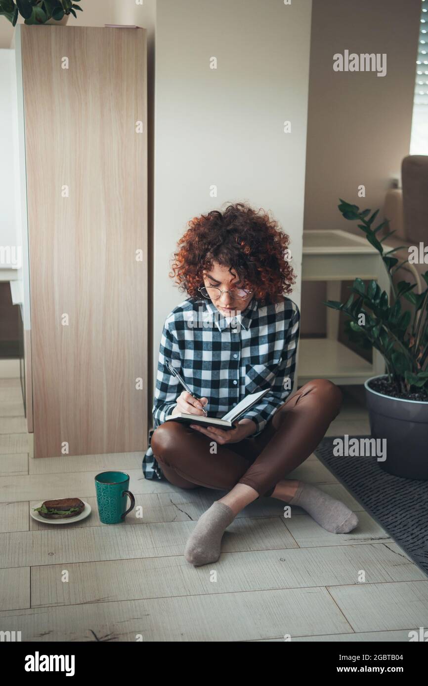 Donna affascinante con capelli ricci sta scrivendo qualcosa in un libro mentre lavora e beve un caffè con sandwich Foto Stock