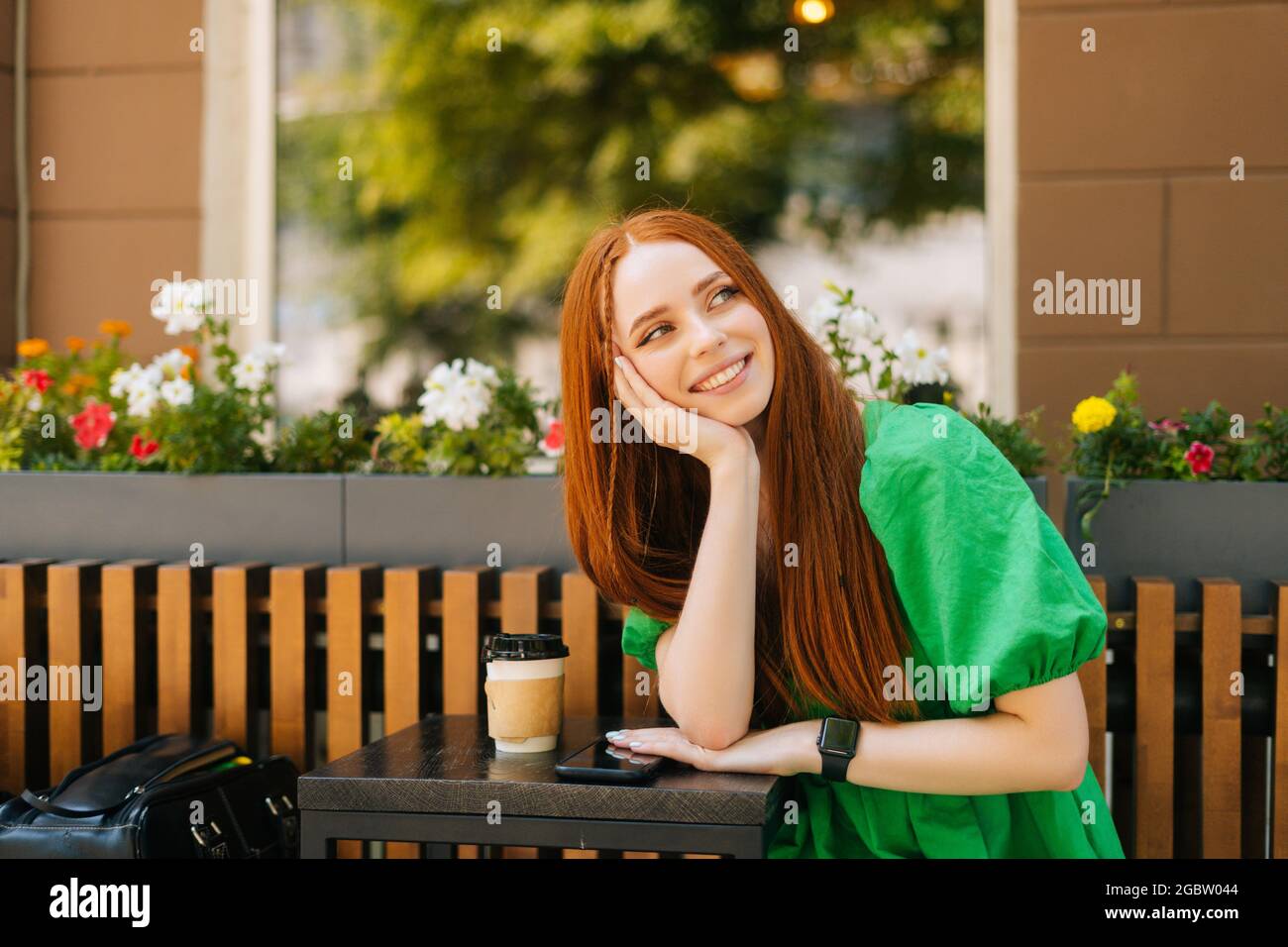 Ritratto di giovane donna con winking rosso, seduto a tavola con caffettiera e telefono cellulare in terrazza all'aperto nella soleggiata giornata estiva Foto Stock