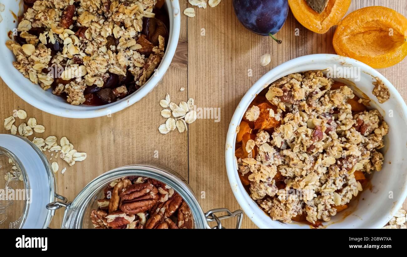 Crumble appena sfornato con albicocca e frutta di prugne, noci di pecan e avena arrotolata su tavola di legno. Un vaso di vetro con noci pecan aperte. Vista dall'alto, primo piano. Foto Stock