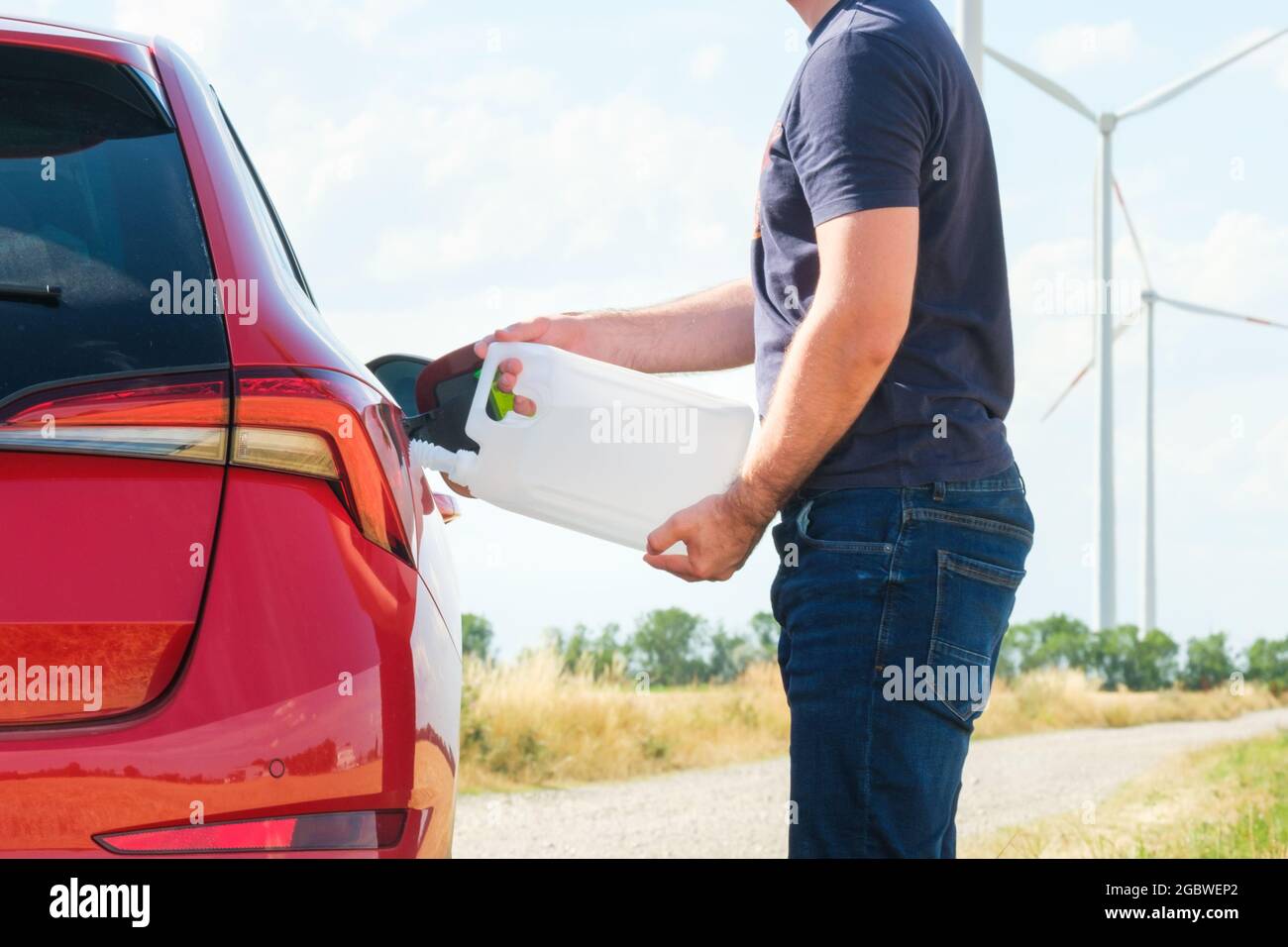 Uomo che riempie un liquido motore diesel dal serbatoio nel serbatoio dell'auto rossa sullo sfondo delle turbine eoliche. Riduzione dell'inquinamento atmosferico e. Foto Stock
