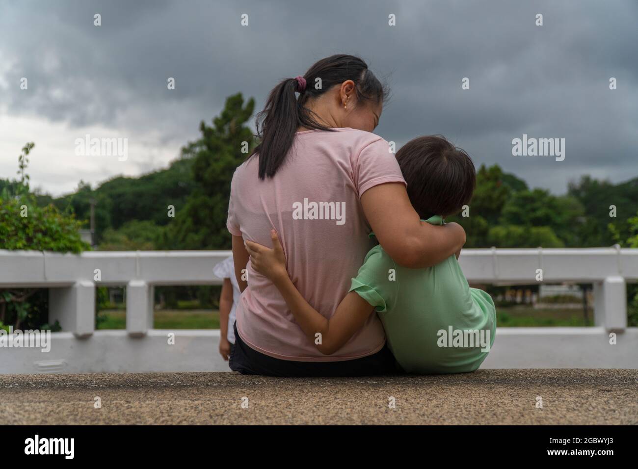 Vista posteriore di amorevole madre asiatica abbraccio i suoi bambini seduti sul ponte, la mamma nera cura abbraccio bambino, rilassante guardando alla nuvola nera, genitore conforto bambino Foto Stock