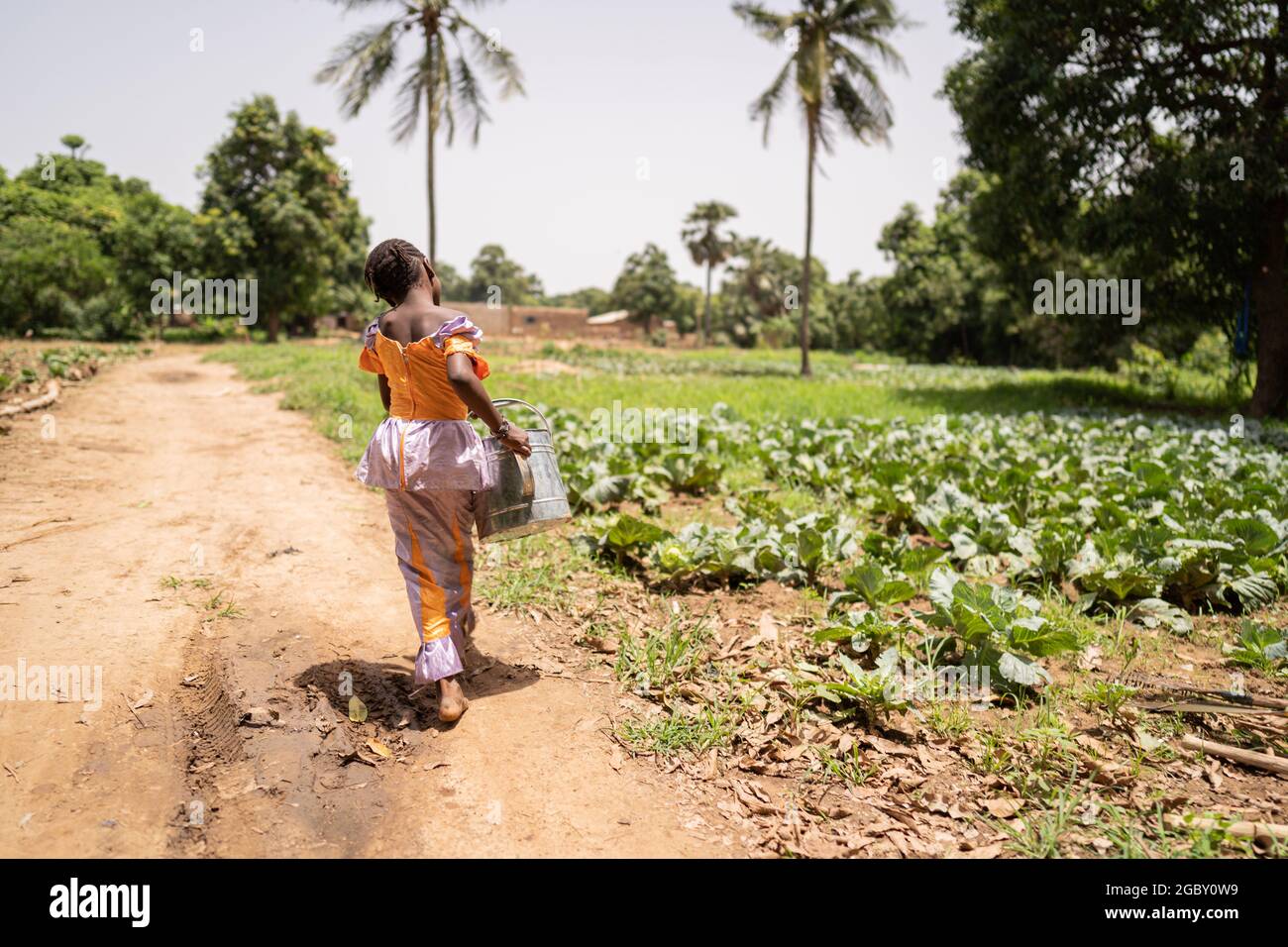 Piccola ragazza africana nera lotta per portare una lattina di irrigazione pesante dal pozzo al campo; concetto di lavoro infantile Foto Stock