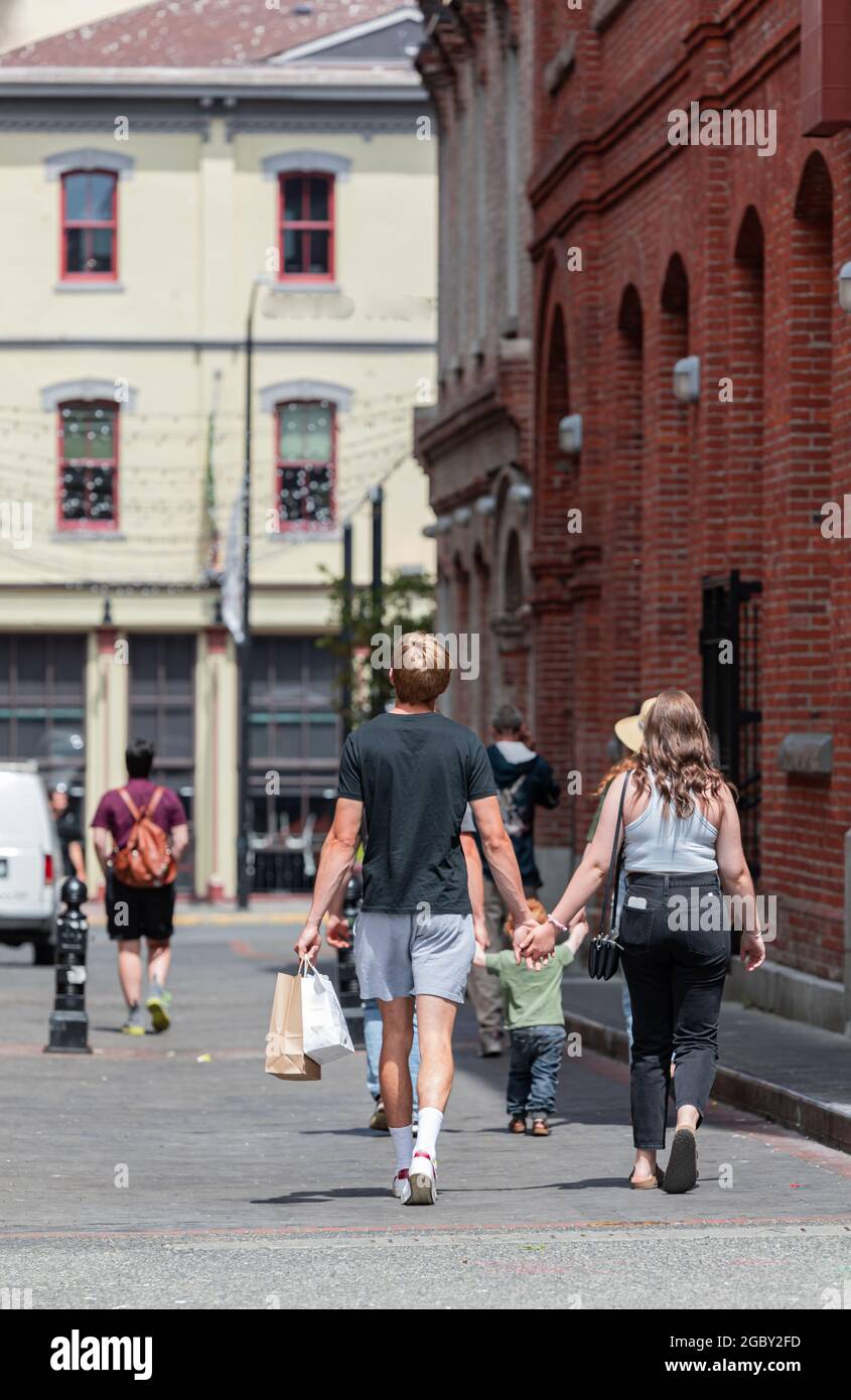 La vista posteriore di una giovane coppia che cammina lungo la strada tenendo le mani e borse shopping. Victoria BC, Canada-Luglio 23,2021. Vista sulla strada, foto di viaggio, Foto Stock