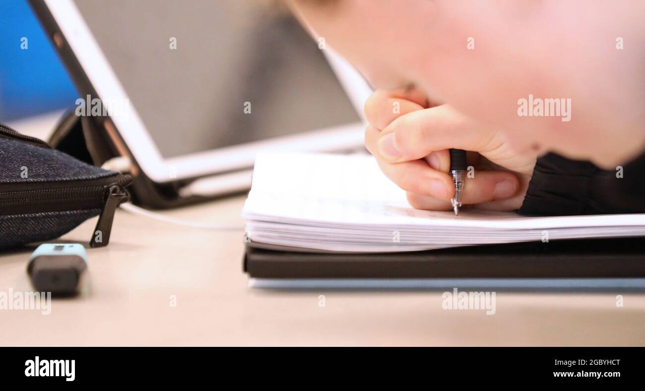 Un primo piano di uno studente che scrive con una penna in un libro di scuola a una scrivania. Custodia a matita e cancellatore visibili con un dispositivo digitale o un tablet nel retro Foto Stock