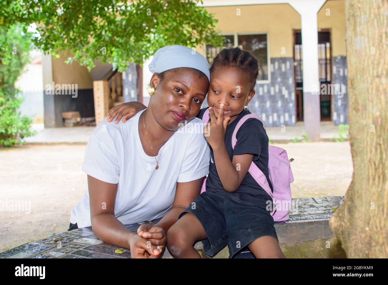 Madre africana o tutore seduto insieme con la sua bambina sotto un albero in un ambiente scolastico, disponibile per aiutare la figlia eccellenza in lei Foto Stock