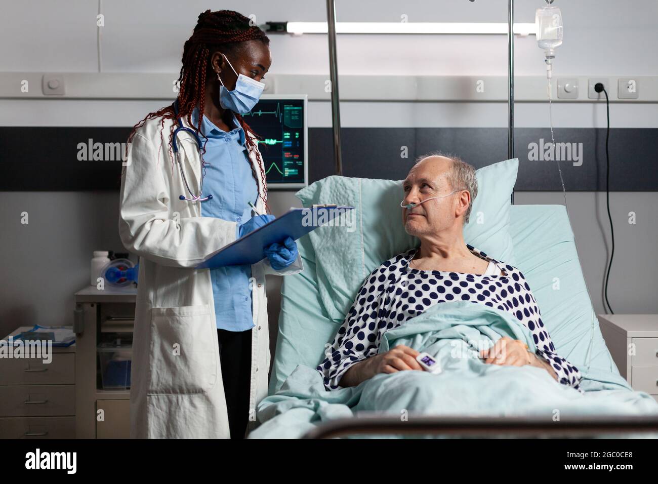 Medico afroamericano con maschera chirurgica che legge la diagnosi dalla clipboard a malato malato malato paziente anziano che si posa a letto, respirando con l'aiuto della maschera di ossigeno, ascoltando discutere con il personale medico circa il recupero. Foto Stock