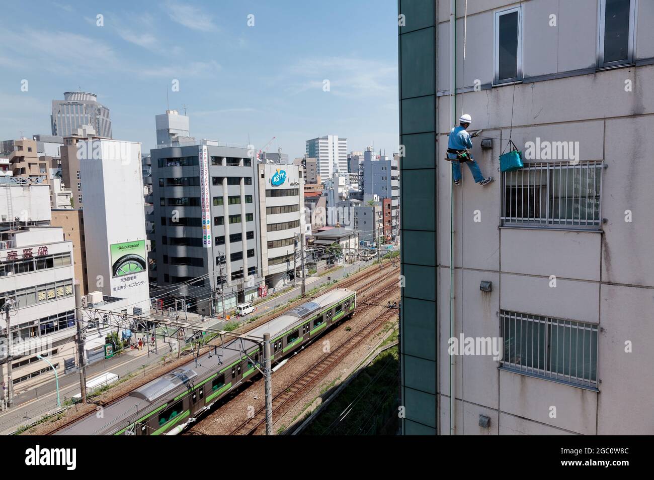 Un tecnico addetto all'accesso alle funi ripara un edificio di appartamenti mentre un treno della linea Yamanote passa sotto. Ebisu, Tokyo, Giappone. Foto Stock
