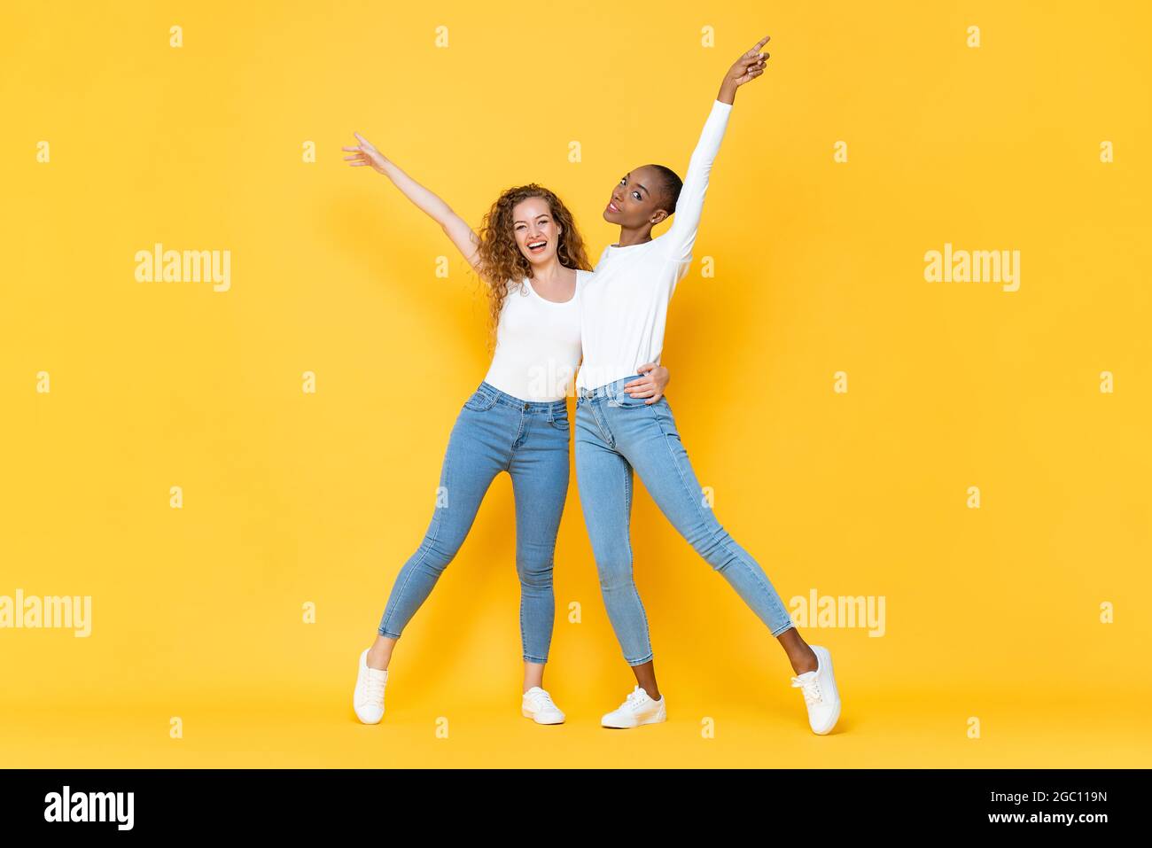 Ritratto a lunghezza intera di sorridere due amici interrazziali millenni donna che si tengono e alzando le mani in su isolato studio giallo colore backg Foto Stock