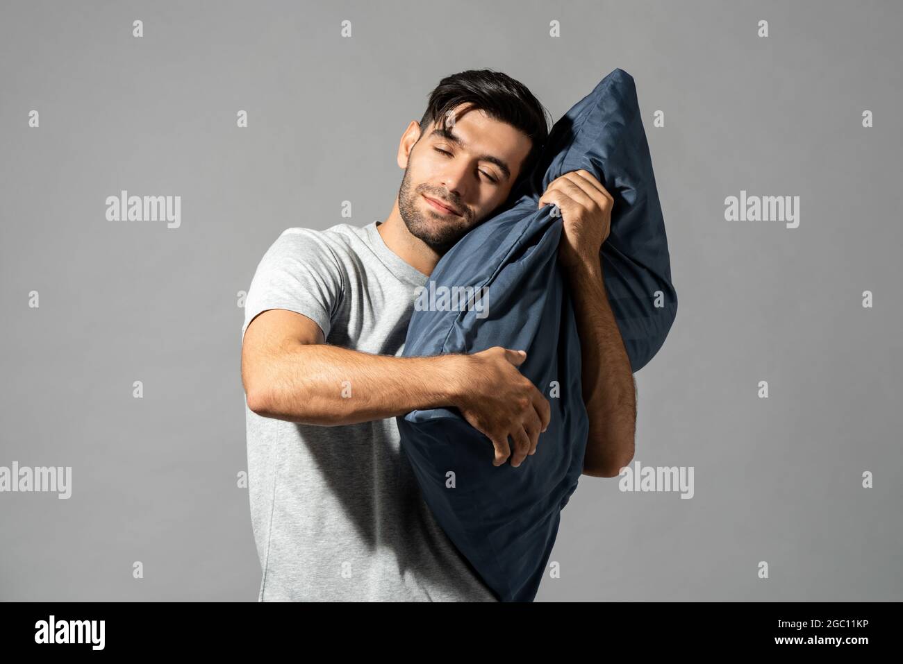 Ritratto isolato di un giovane sorridente che tiene un cuscino chiudendo gli occhi e sognando su uno sfondo grigio chiaro dello studio Foto Stock