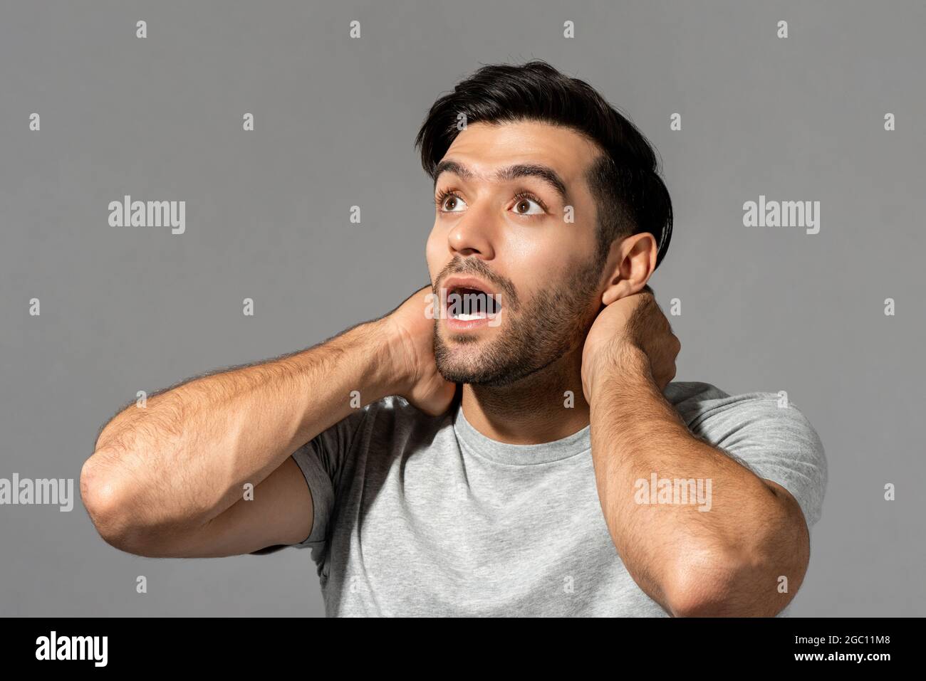 Stordito giovane uomo caucasico gasting con le mani intorno al collo guardando verso l'alto su sfondo grigio chiaro studio Foto Stock