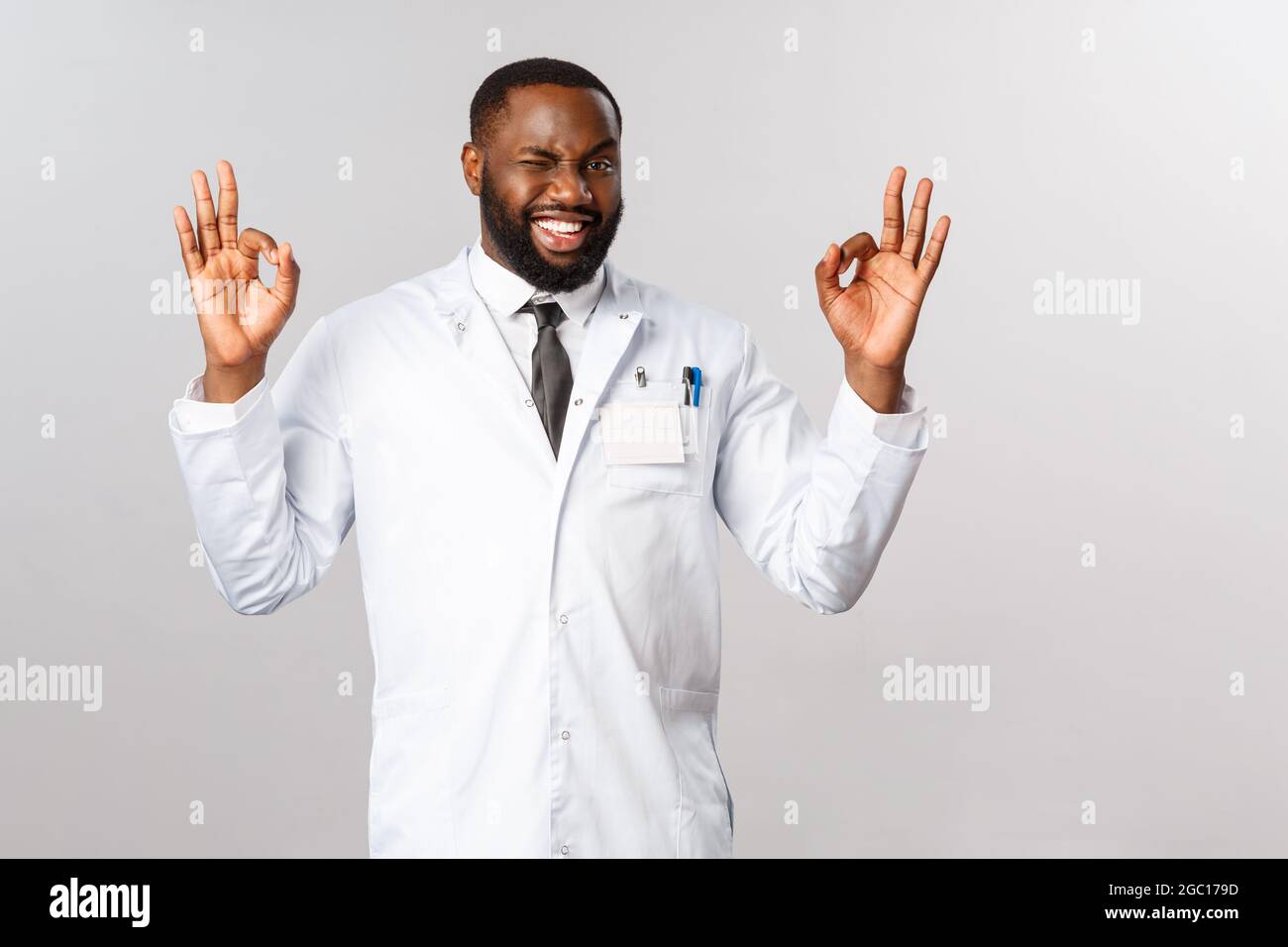 Nessun problema, clinica garantire il miglior servizio professionale e la cura. Medico maschio afroamericano facile-andando in cappotto bianco, inocchiato cheeky e mostrare okay Foto Stock