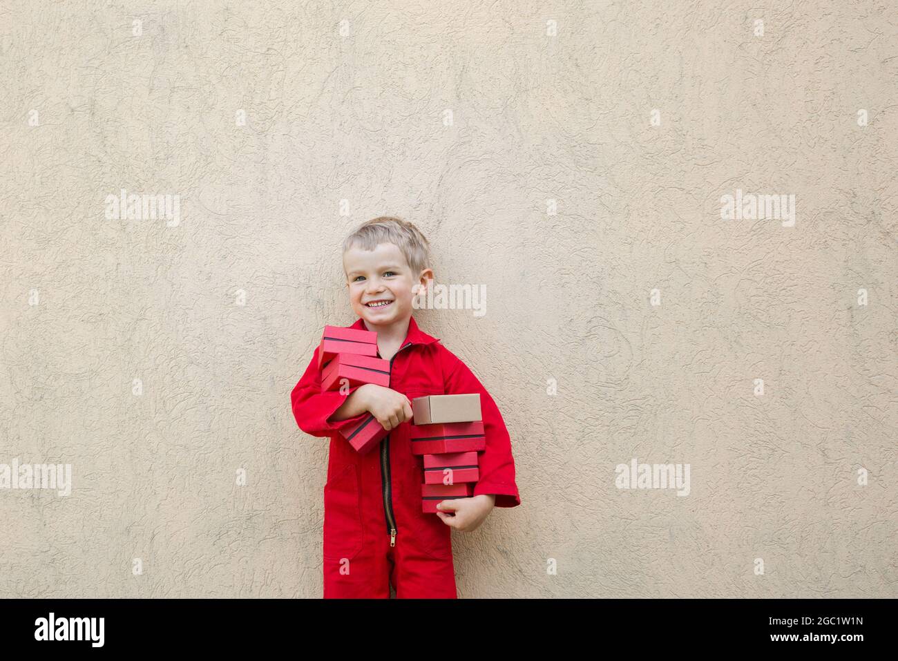 Sorridente simpatico bambino in una tuta rossa mantiene molte piccole scatole nelle mani. Consegna pacchi, piccolo postino, assistente. Positivo, gioia, gam Foto Stock