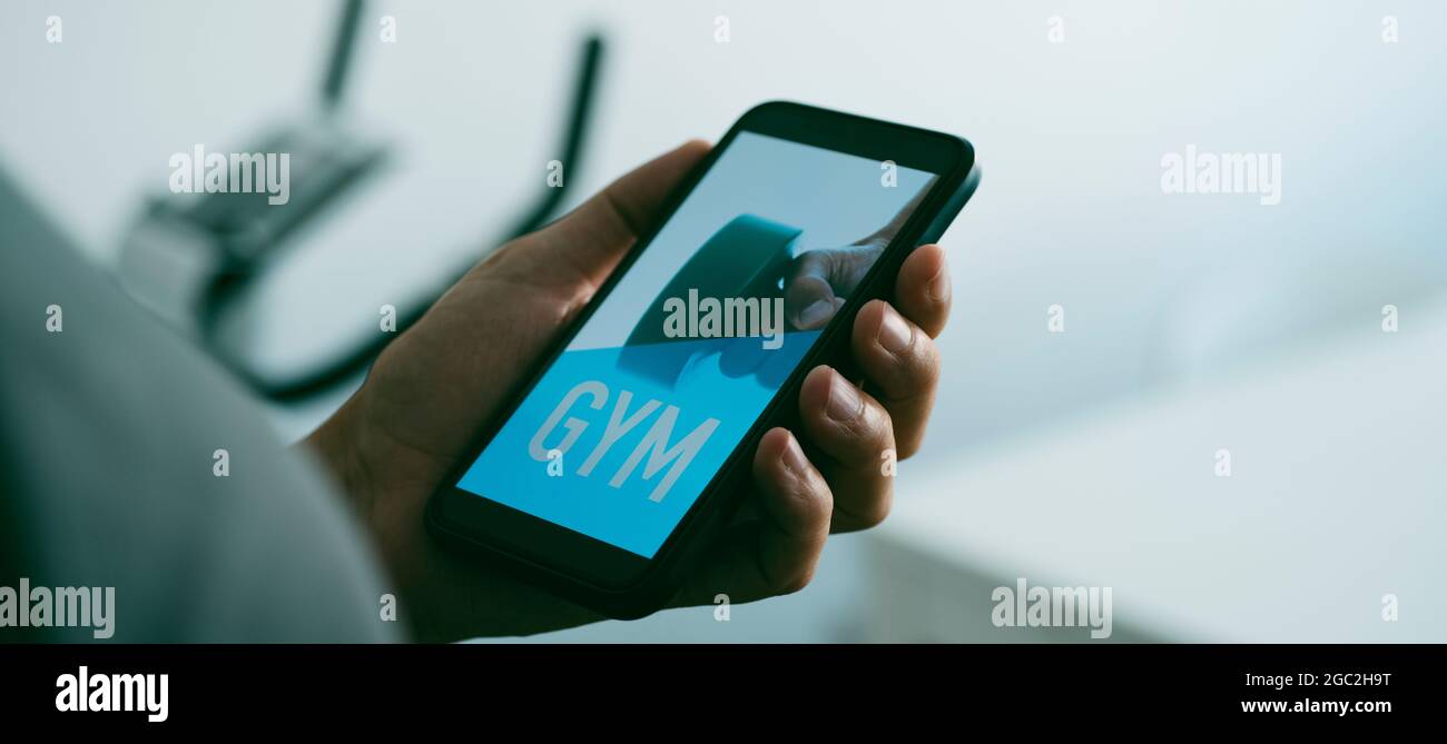 un giovane caucasico sta guardando al suo smartphone, che legge la palestra nel suo schermo, accanto ad una bicicletta stazionaria, in un formato panoramico da utilizzare come web ba Foto Stock