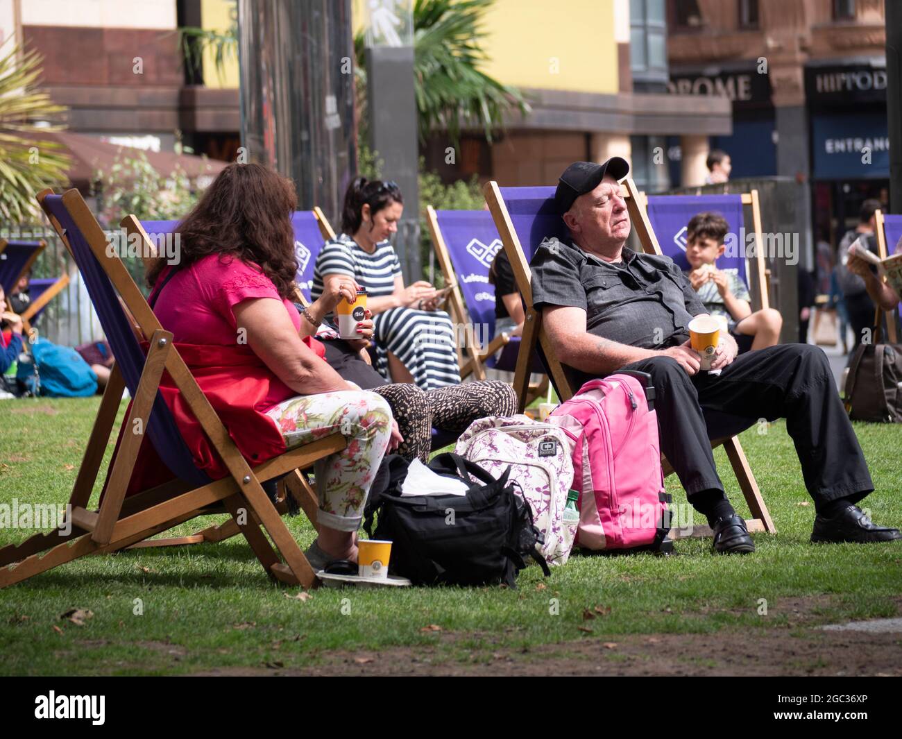 I turisti e gli impiegati che si siedono su sedie a sdraio in erba al sole estivo in piazza Leicester, durante l'ora di pranzo Foto Stock