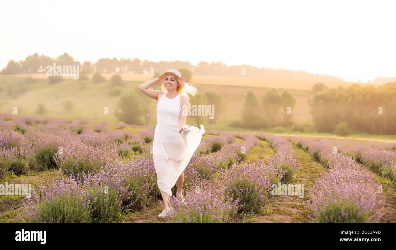 Bella giovane donna sana con un vestito bianco che corre gioiosamente attraverso un campo di lavanda, cappello di paglia Foto Stock