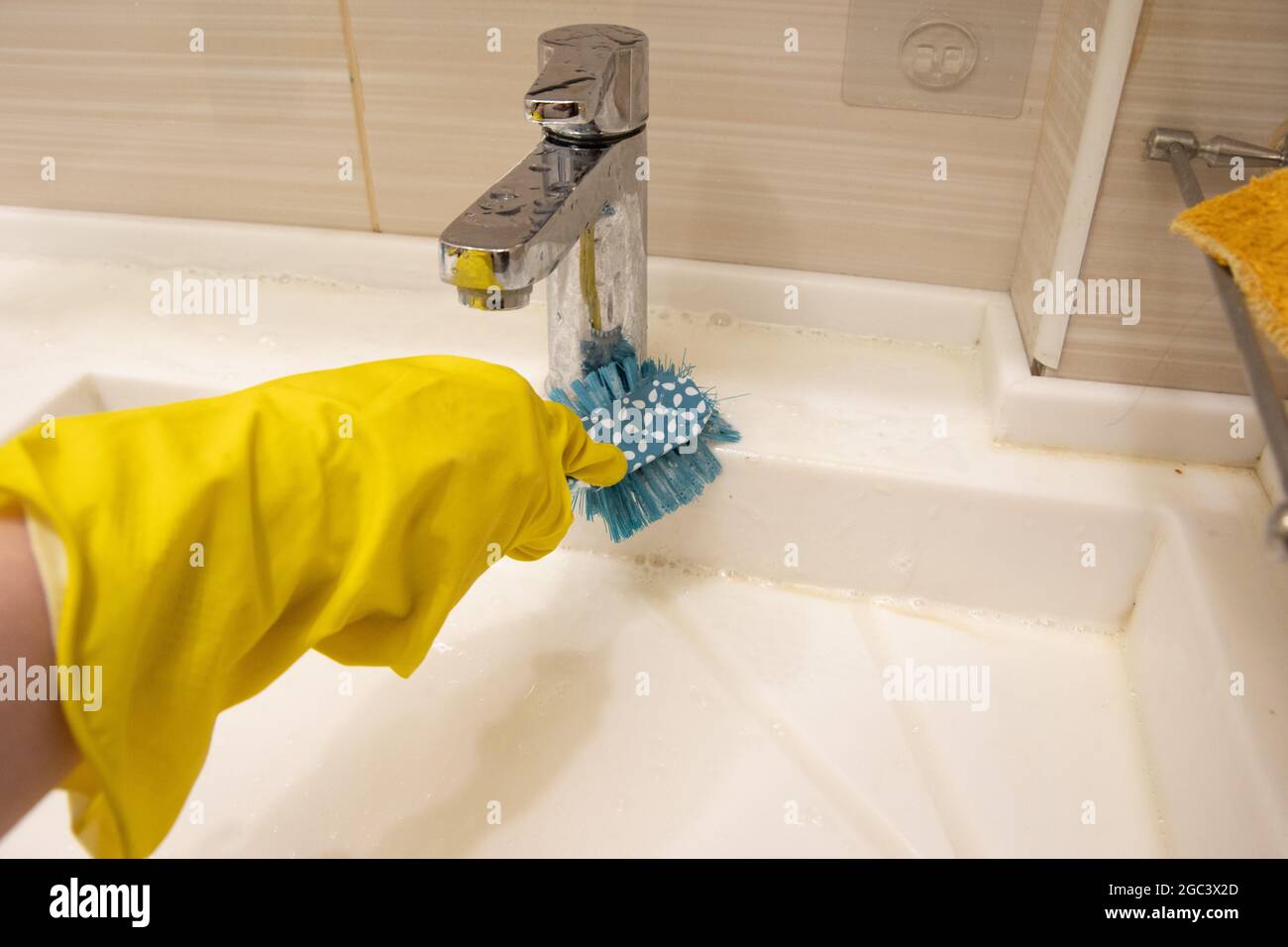 Una mano in un guanto giallo pulisce il lavandino con una spazzola blu sull'impugnatura. Il concetto di lavoro a casa, disinfezione. Foto Stock