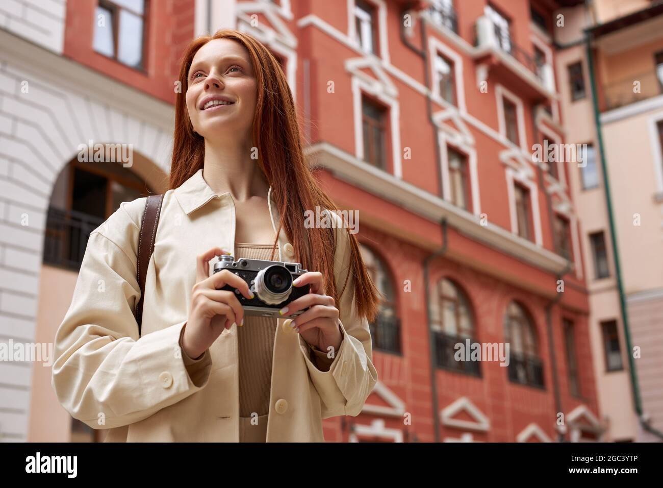 Donna fotografo redhead con fotocamera retrò in mano guardando la vecchia architettura urbana, scattando foto, scoprire nuovi luoghi, camminare da solo in città. Foto Stock