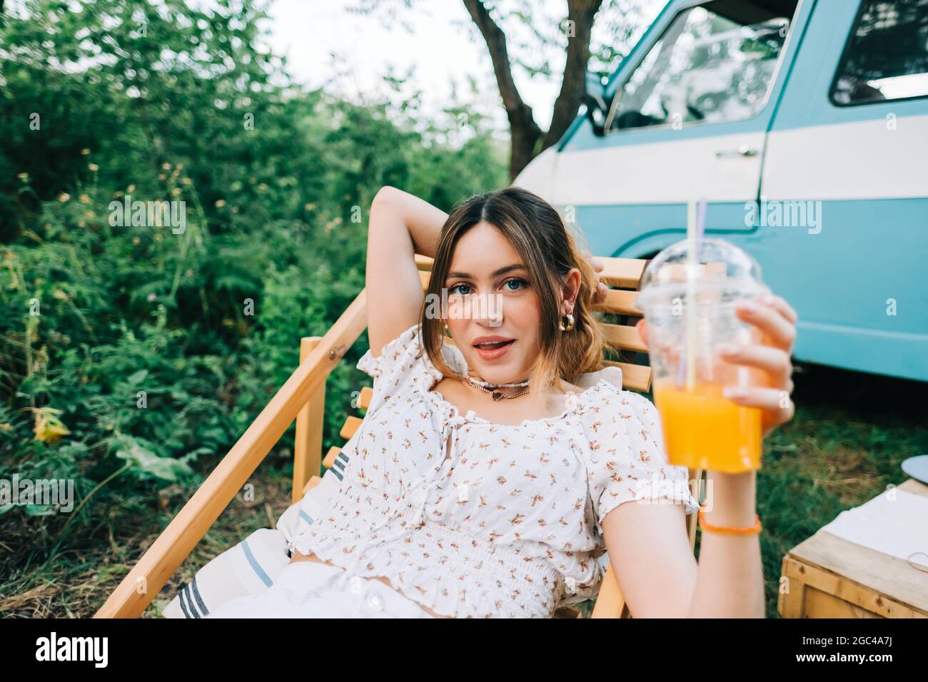 Ritratto di giovane donna seduta su sedia di legno vicino furgone e bevendo limonata all'aperto in natura. Goditi l'estate, il concetto di viaggio Foto Stock