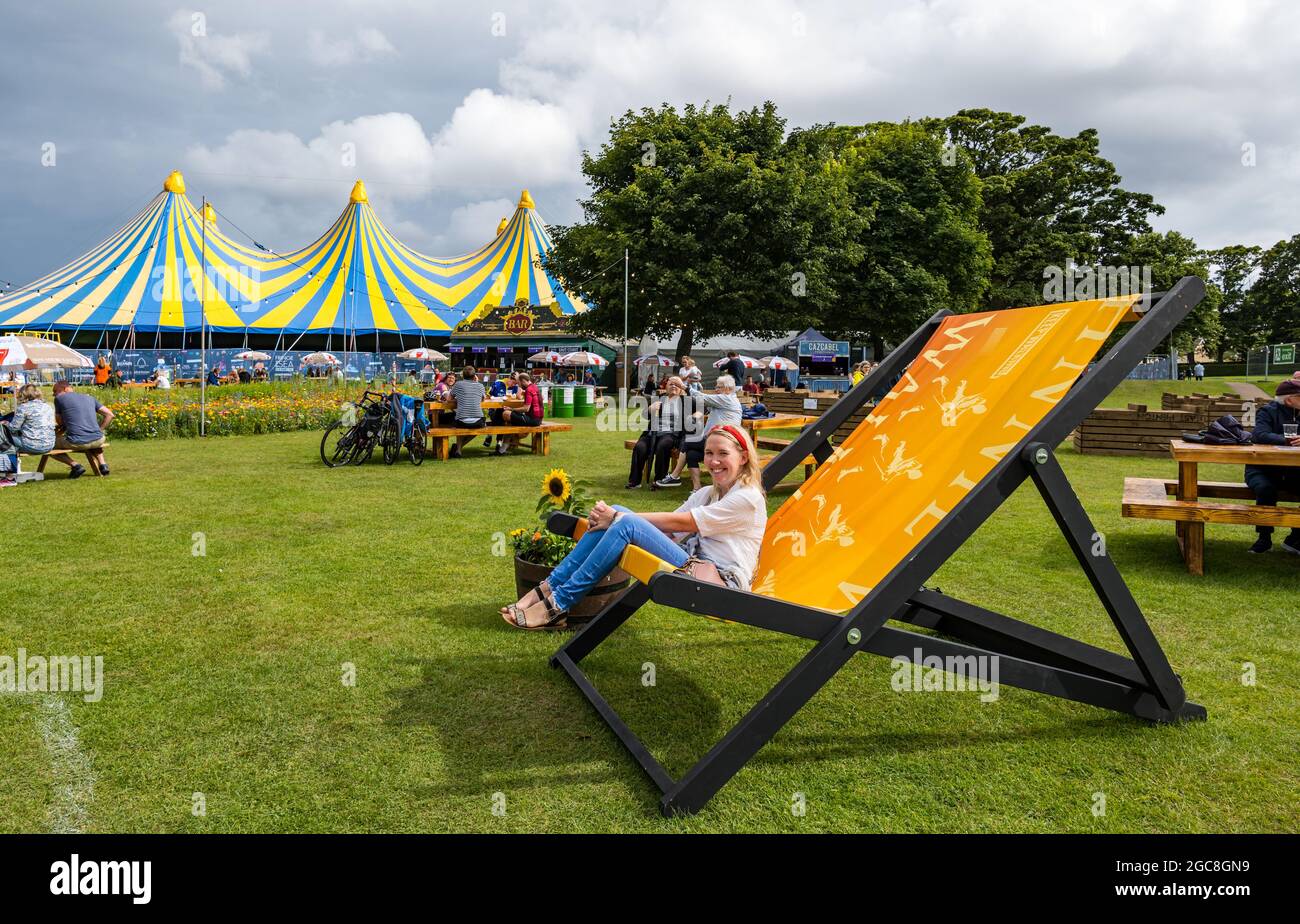 North Berwick, East Lothian, Scozia, Regno Unito, 7 agosto 2021. Regno Unito Meteo: Sole e docce a Fringe-by-the-Sea giorno 2. Nella foto: Una donna si siede su una sedia gigante nel luogo principale del festival nel parco Lodge Grounds, vicino alla tenda Belhaven Big Top Foto Stock