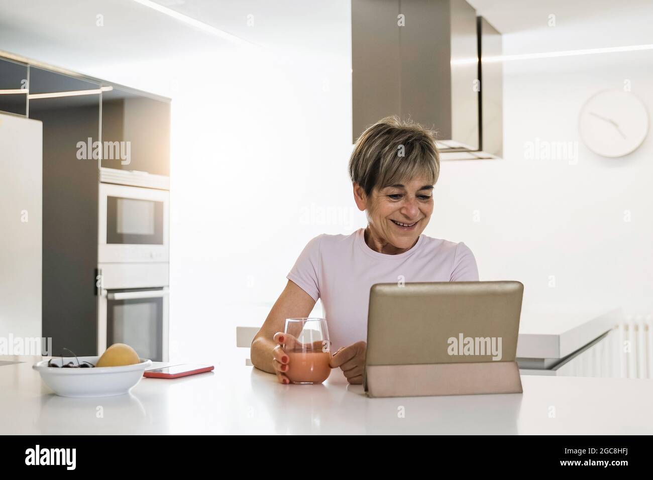 Donna anziana felice che usa il tablet digitale mentre si mangia la colazione in cucina - Focus on Face Foto Stock