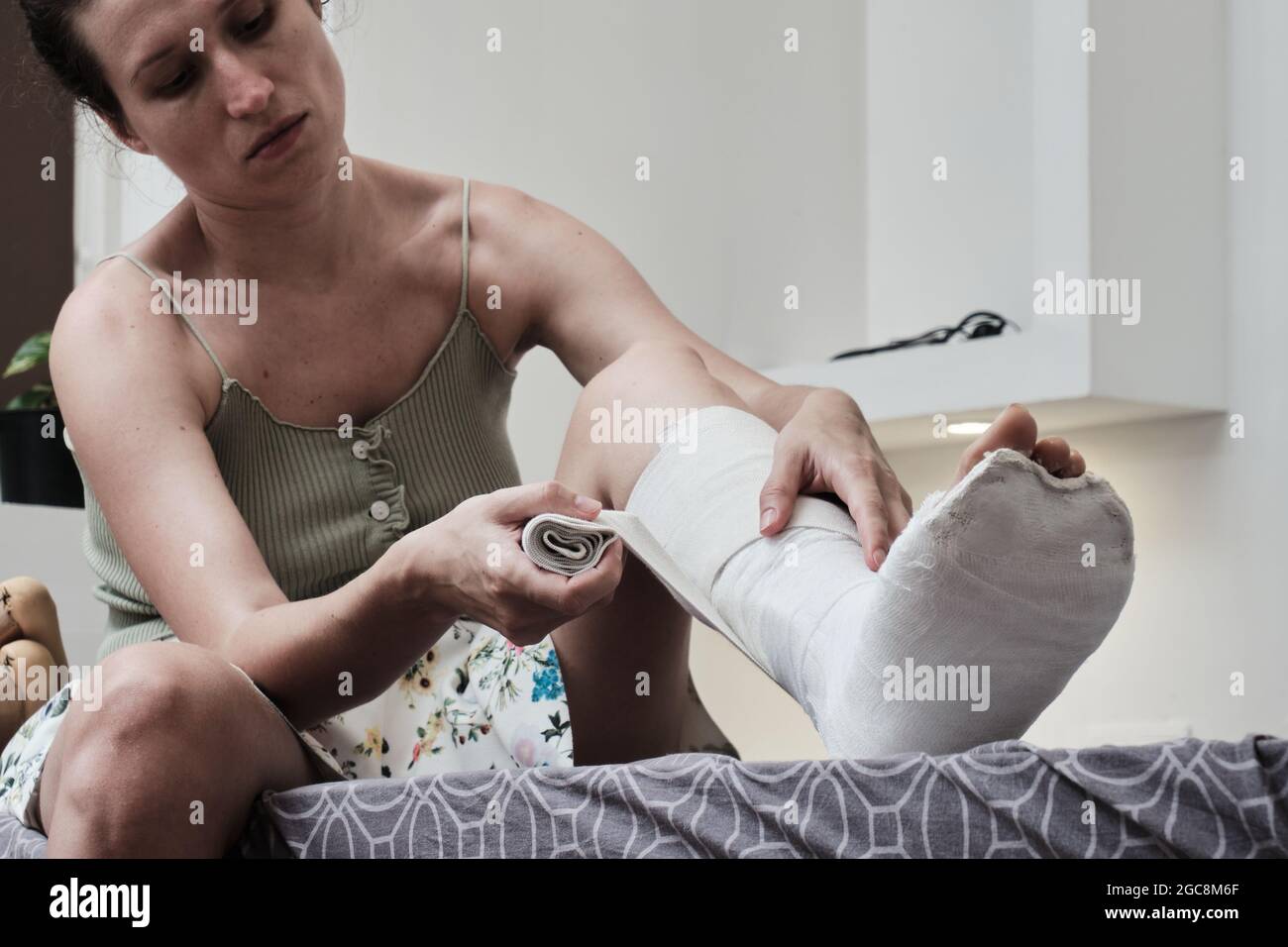 La donna seduta su un letto mette un bendaggio elastico su una gamba rotta. La gamba è in un calco di gesso. riabilitazione domestica dopo una gamba rotta. Pho di alta qualità Foto Stock