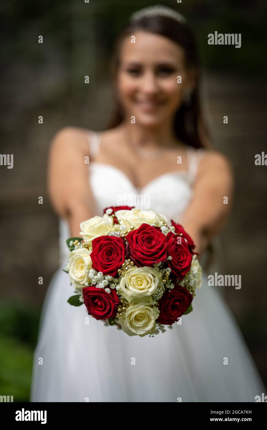 Symbole, Feature, Deutschland, Eine Braut (im Hintergrund unscharf) in weißem Brautkleid zeigt ihren Brautstrauß der aus gelben, weißen und roten Rose Foto Stock