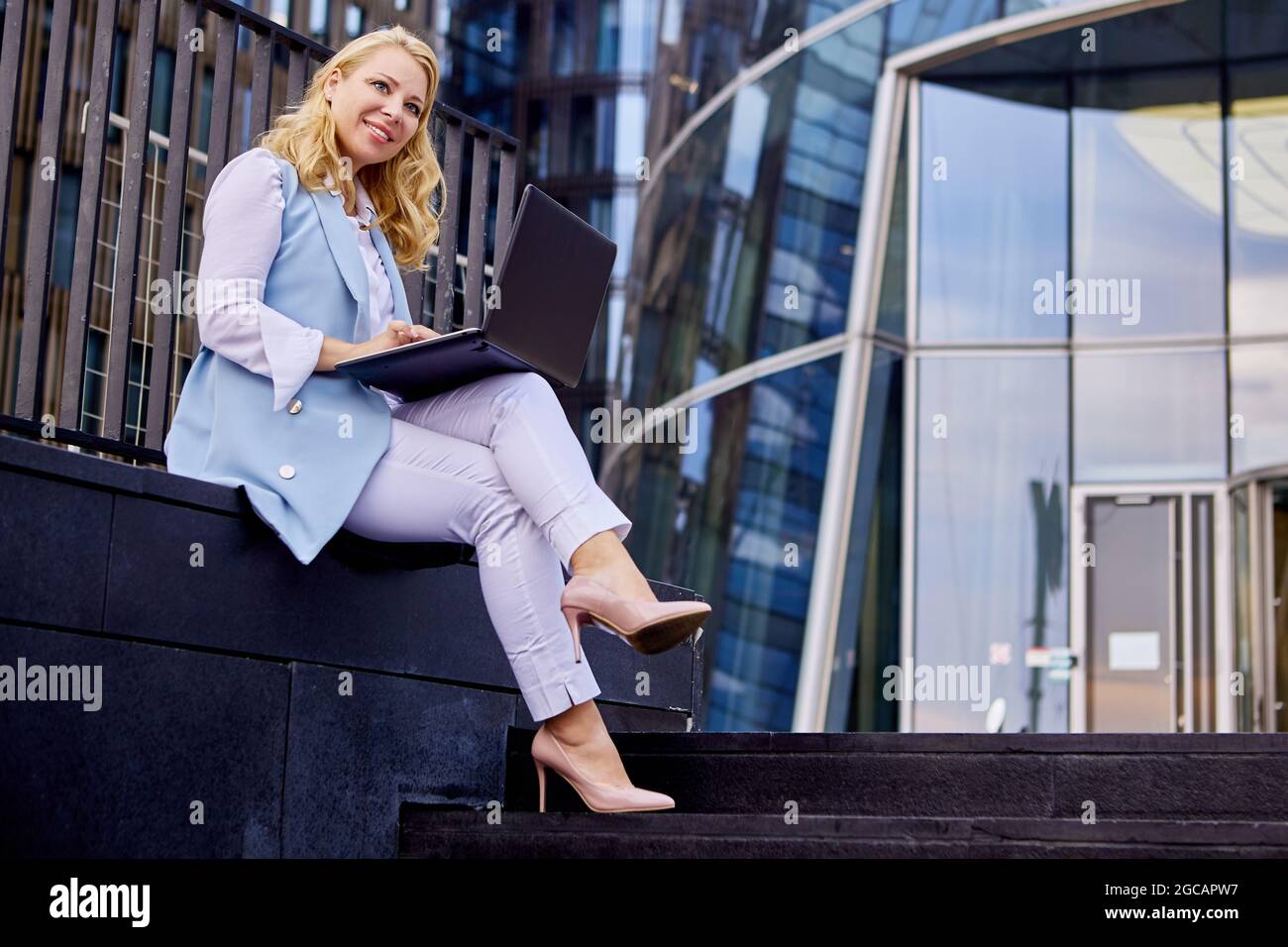Una donna sorridente in tuta da lavoro con computer portatile è seduta all'aperto. Foto Stock