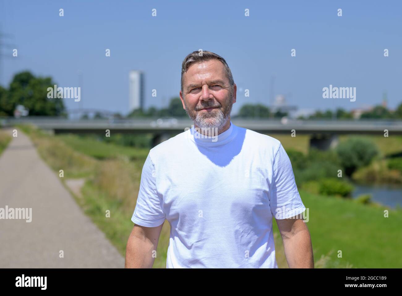 Uomo bearded di mezza età che sorride alla telecamera mentre fa jogging su un sentiero accanto ad un fiume in una bella giornata estiva soleggiata in un concetto di un AC sano Foto Stock