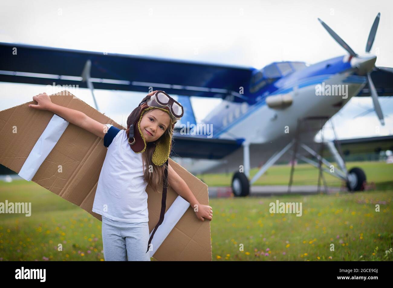 Una bambina in costume da pilota con ali di cartone corre sul prato contro lo sfondo dell'aereo. Un bambino in un cappello e gli occhiali sogna Foto Stock