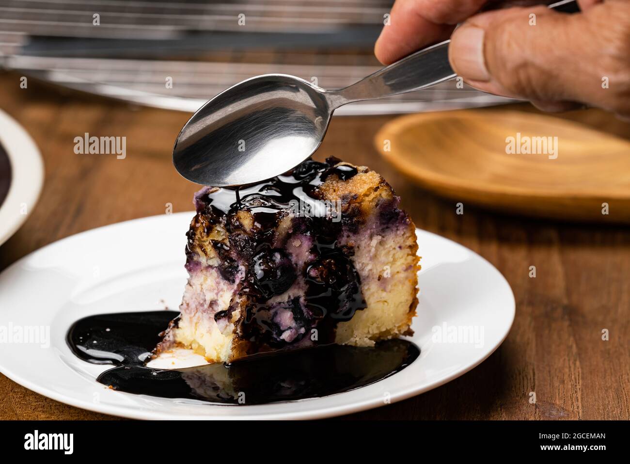 Mano più anziana con cucchiaio di metallo mangiare delizioso mirtillo fatto in casa e crumble cheesecake condimento con cioccolato in piastra di ceramica bianca su tavolo di legno Foto Stock