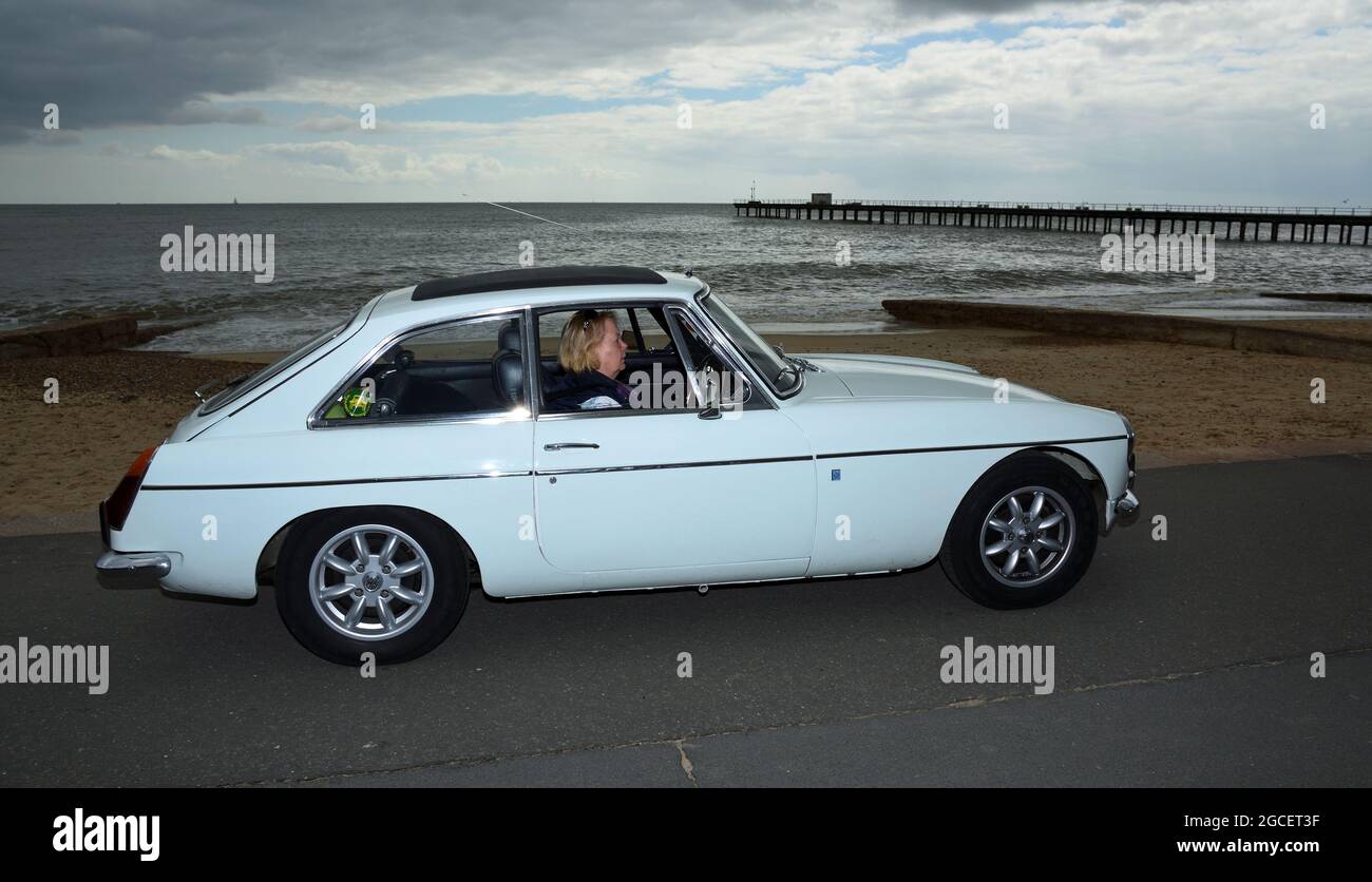 La classica Auto White MGB GT è guidata lungo la passeggiata lungomare con cielo tempestoso. Foto Stock