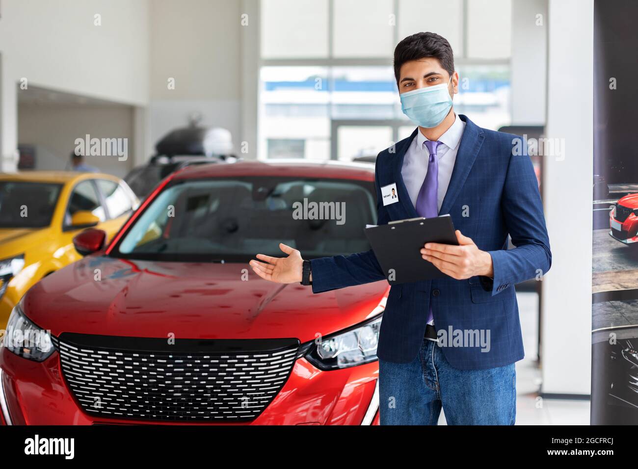 Uomo arabo in abito formale e maschera protettiva in piedi su auto rossa di lusso in grande showroom, copia spazio. Responsabile vendite maschile che punta a nuovo di zecca Foto Stock