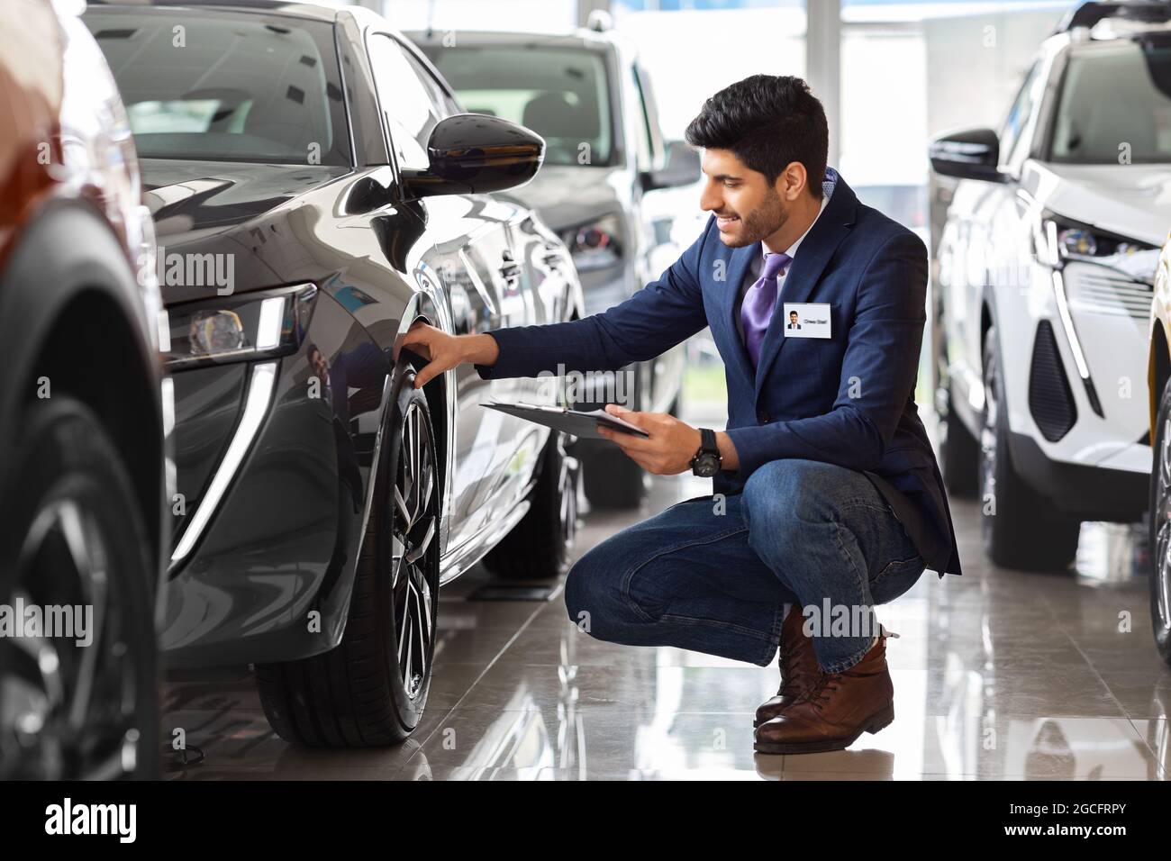 Bell'uomo in tuta assistente di vendita controllo pneumatici auto presso showroom di lusso, con piegatrice con documenti. Medio-orientale Guy responsabile vendite in salone auto Foto Stock