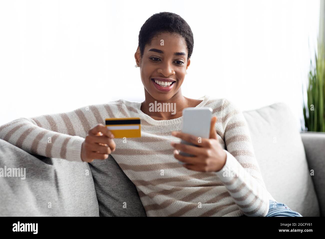 Young Black Lady con smartphone e carta di credito a casa utilizzando l'app Online Banking mentre si riposa sul CouchIn soggiorno, Happy African Amerian Woman SH Foto Stock