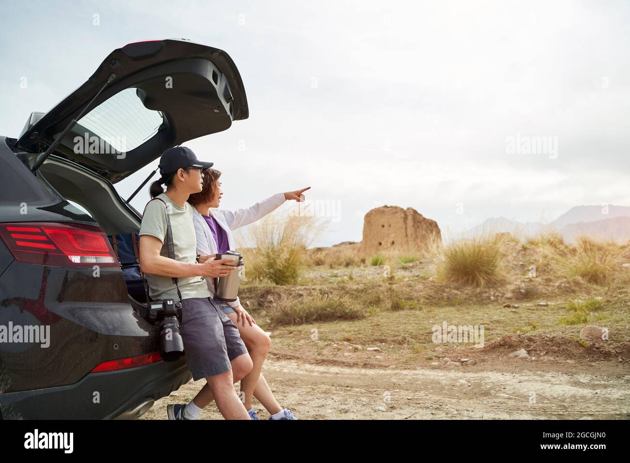 la coppia asiatica si appoggia contro il retro dell'auto guardando la vista mentre si beve caffè in un sito storico desolato Foto Stock