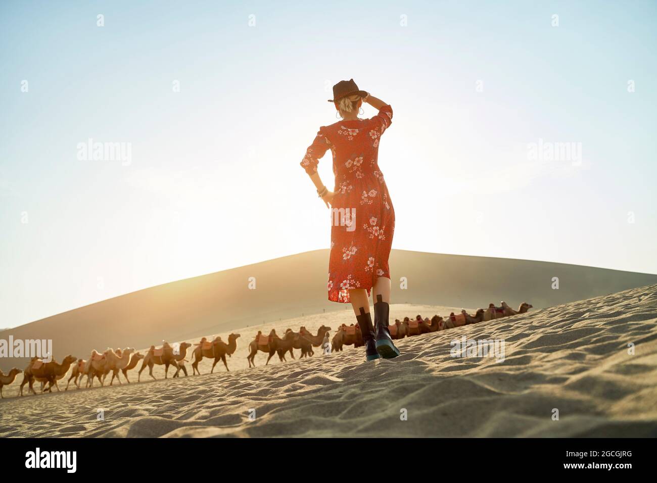 vista posteriore della donna asiatica in abito rosso in piedi nel deserto guardando la vista con caravan di cammelli e enorme duna di sabbia sullo sfondo Foto Stock